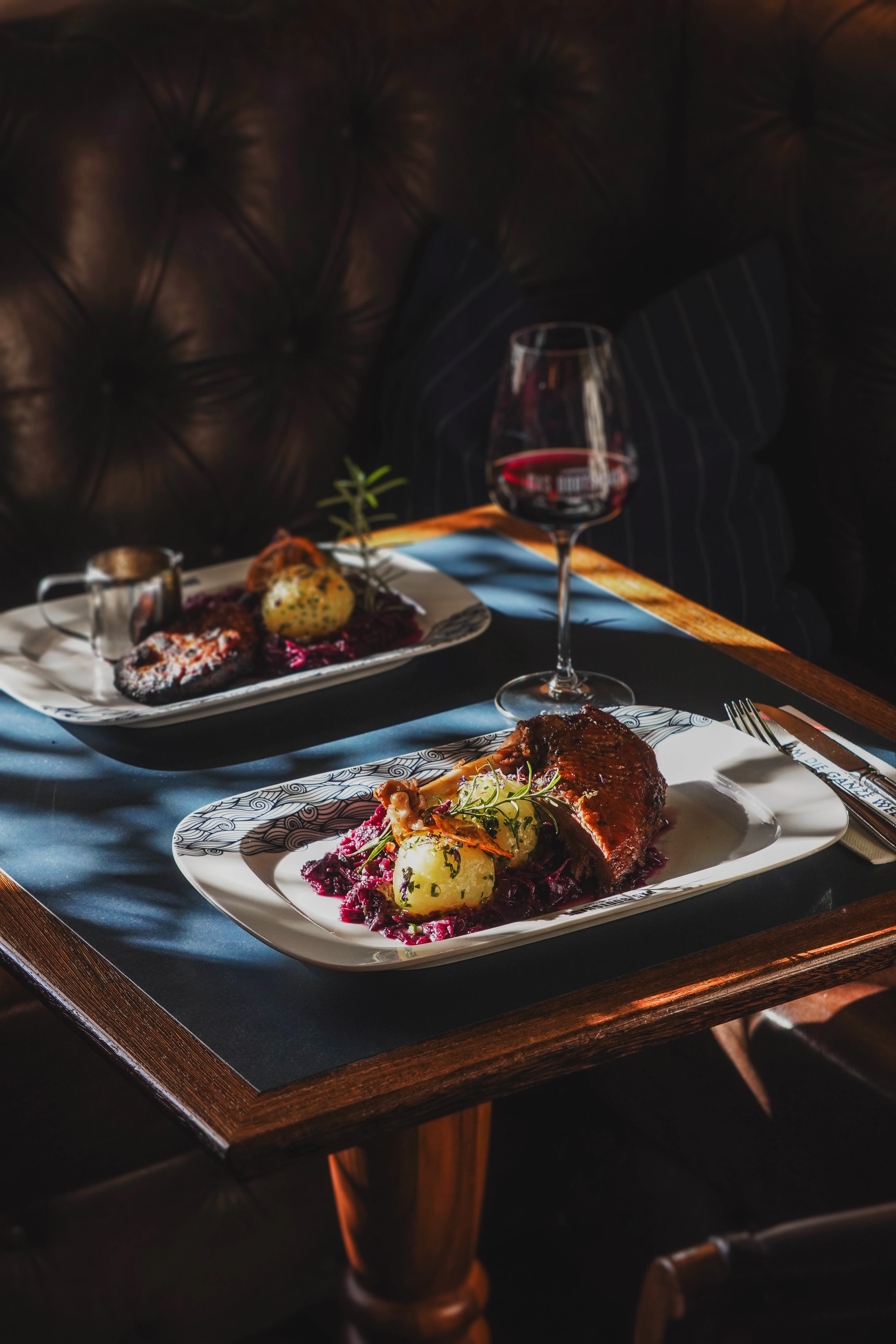 Zwei Teller mit traditionellem Gänsebraten, Rotkohl und Knödeln auf einem Holztisch in einem Restaurant, begleitet von einem Glas Rotwein.